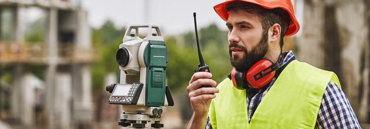 A land surveyor in hard hat and safety vest speaks into a walkie talkie