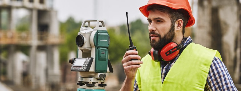 A land surveyor in hard hat and safety vest speaks into a walkie talkie