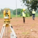 A yellow surveying instrument on a tripod in a field, with two workers in reflective vests and helmets in the background, symbolizing precision and teamwork.