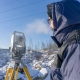A person in a blue winter coat uses surveying equipment on a snowy landscape. Their breath is visible in the cold air, under a clear blue sky.