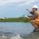 A person in a hard hat and high-visibility vest crouches by the waterside, collecting a water sample with a test tube. The backdrop includes greenery and a cloudy sky.
