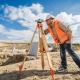 A land survey worker in a hard hat and orange vest uses a theodolite on a tripod at a sunny construction site, with a digger in the background.