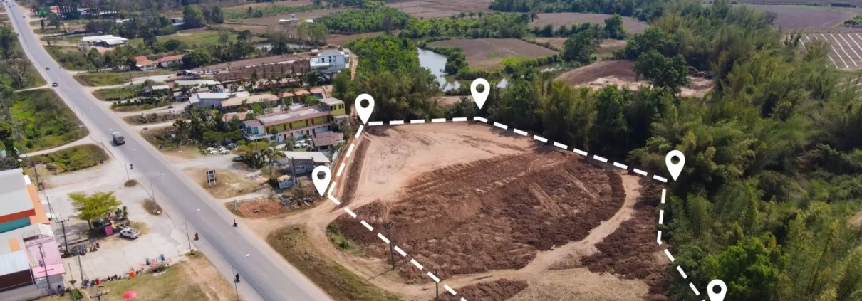 Aerial view of a cleared plot of land outlined with a dashed line and markers. It's beside a road with scattered houses and lush mountains in the background.