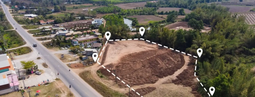 Aerial view of a cleared plot of land outlined with a dashed line and markers. It's beside a road with scattered houses and lush mountains in the background.