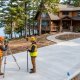 Two land surveyors in high-visibility vests use a total station and prism pole to measure a large, newly poured concrete driveway at a lakeside home in Minnesota.