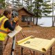 A land surveyor looks through a total station on a tripod beside a lake, with a survey map on a foreground table and a rustic cabin in the background.