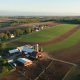 Aerial view of a rural farmstead with red barns, silos, and fenced pastures surrounded by large green and brown crop fields, tree-lined roads, and gently rolling countryside.